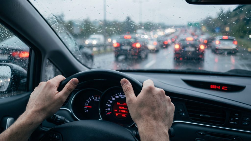 Hands on a steering wheel driving a car in heavy rain with traffic brake lights in the distance