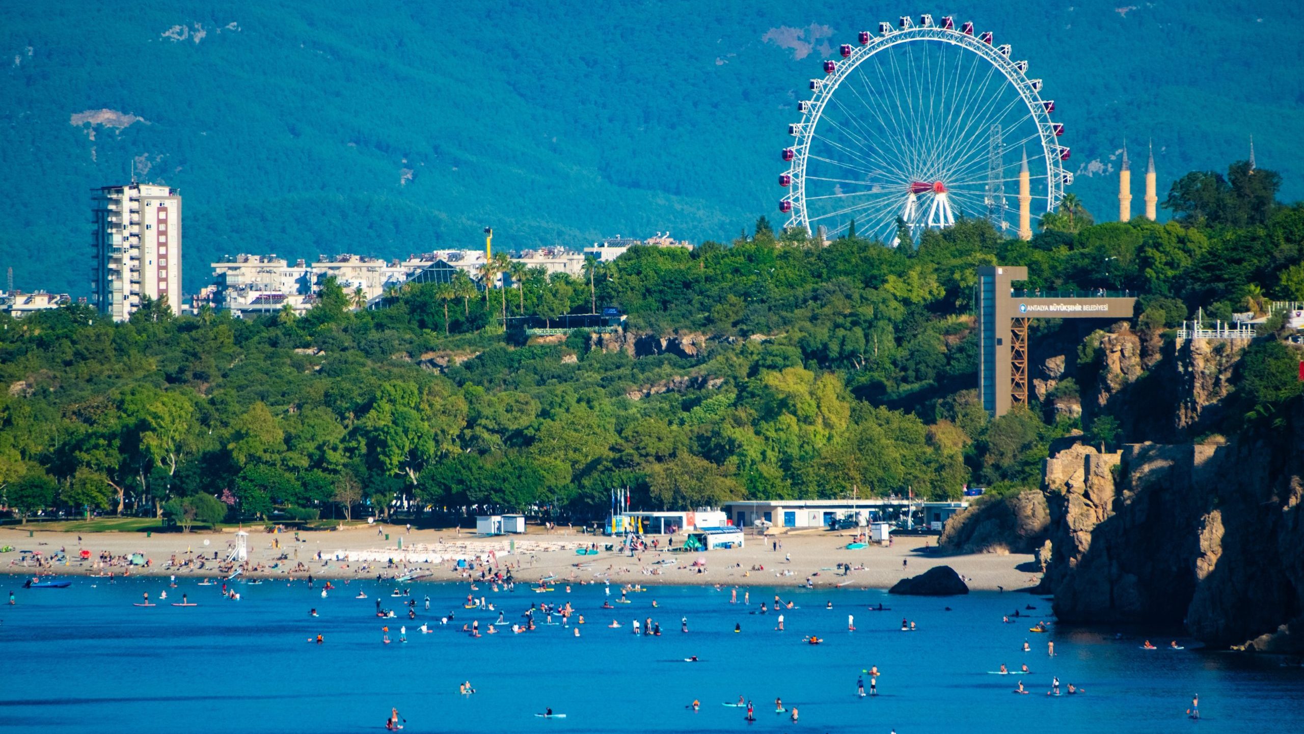 Crowded beach and turquoise sea in Antalya Turkey with a giant Ferris wheel and mountains in the background