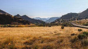 Paved road cutting through a dry golden grassland field with rugged mountains under a clear blue sky in Oregon. (111 characters)