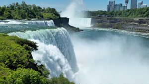 View of American Falls dropping into misty Niagara River gorge with Canadian skyline under clear blue summer sky.
